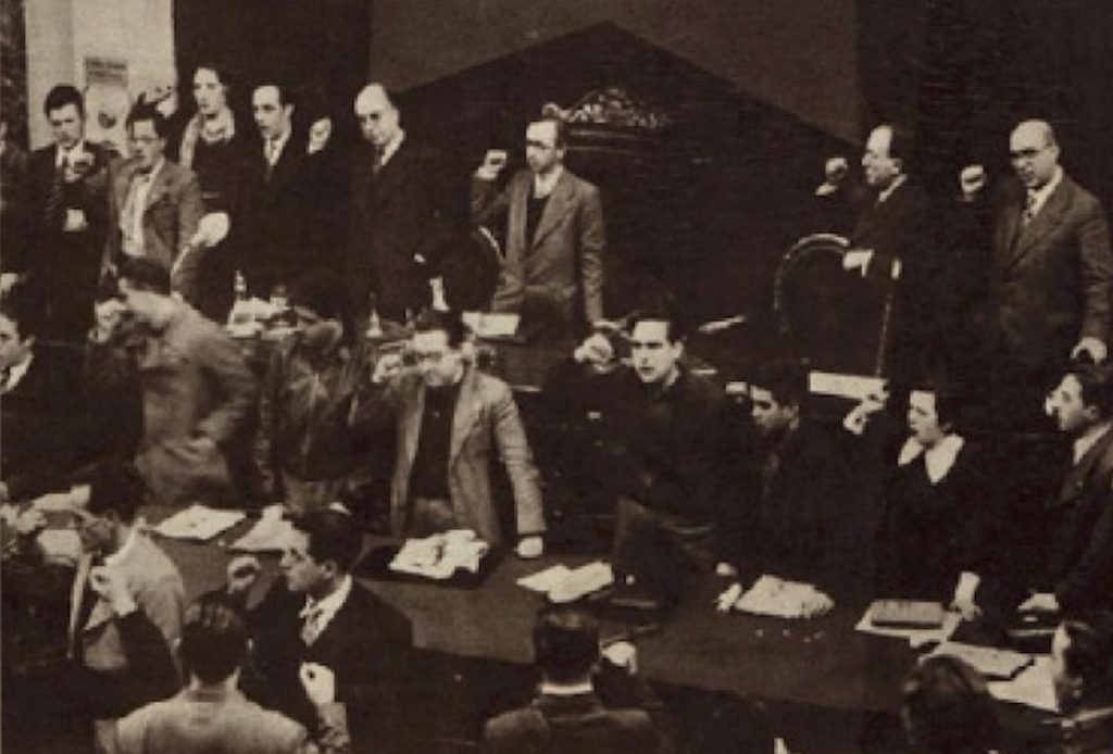 Antonio Machado puño en alto y apoyándose en una silla, el segundo arriba desde la derecha, en el Congreso de las Juventudes Socialistas Unificadas en Valencia en enero de 1937.
